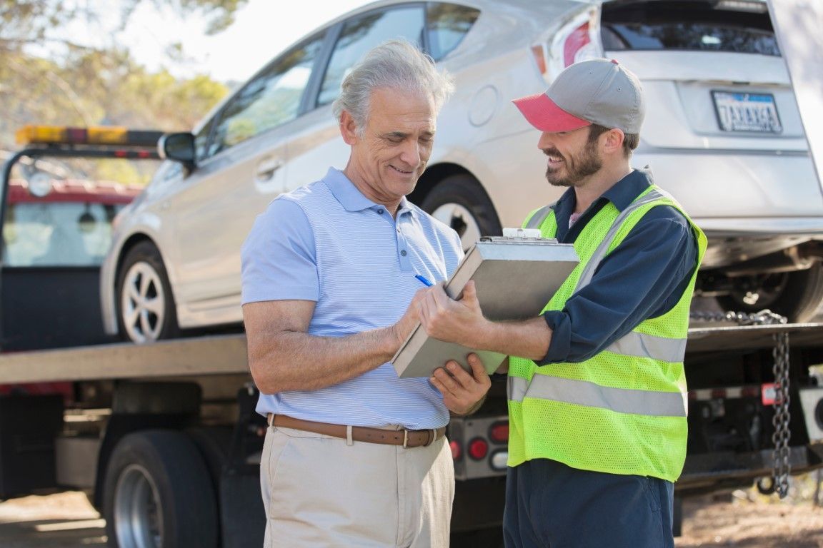 Man signs paperwork with tow truck driver in safety vest next to a white car loaded on a flatbed truck on a sunny day.