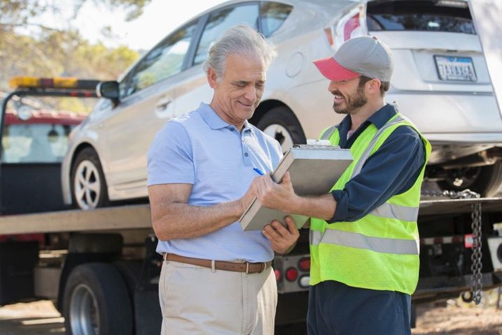Man signs paperwork with tow truck driver in safety vest next to a white car loaded on a flatbed truck on a sunny day.