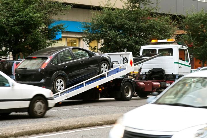 A tow truck lifts a black car in an urban area, likely due to illegal parking or a breakdown, with traffic passing by.