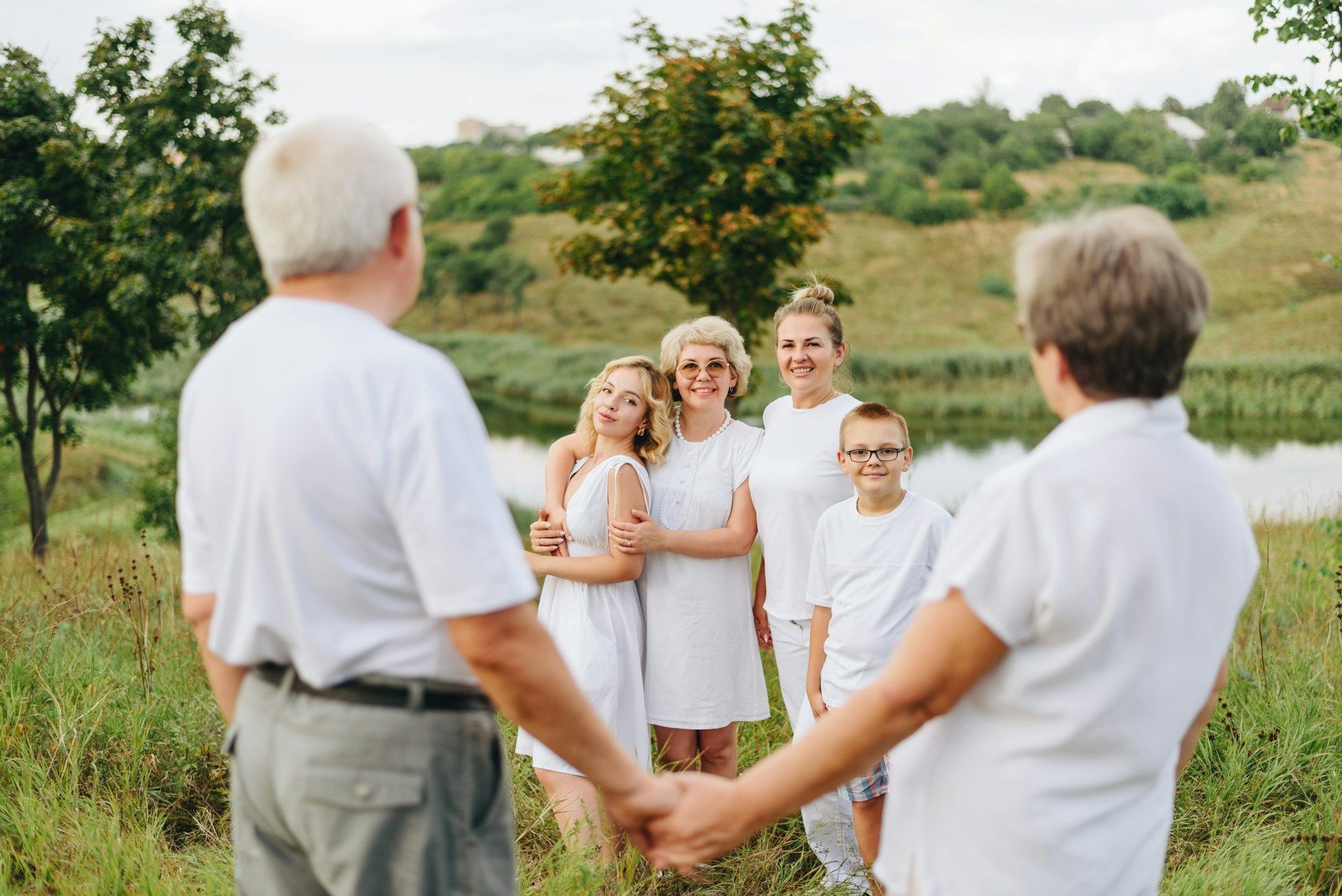 Five people stand hand-in-hand in a grassy field, smiling near a tree-lined hill.