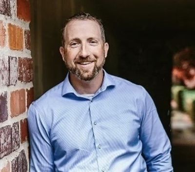 Man in a blue dress shirt leaning against a brick wall, smiling in a dim hallway.