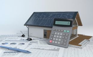 Calculator tilted on top of a cardboard cottage house with solar panels.