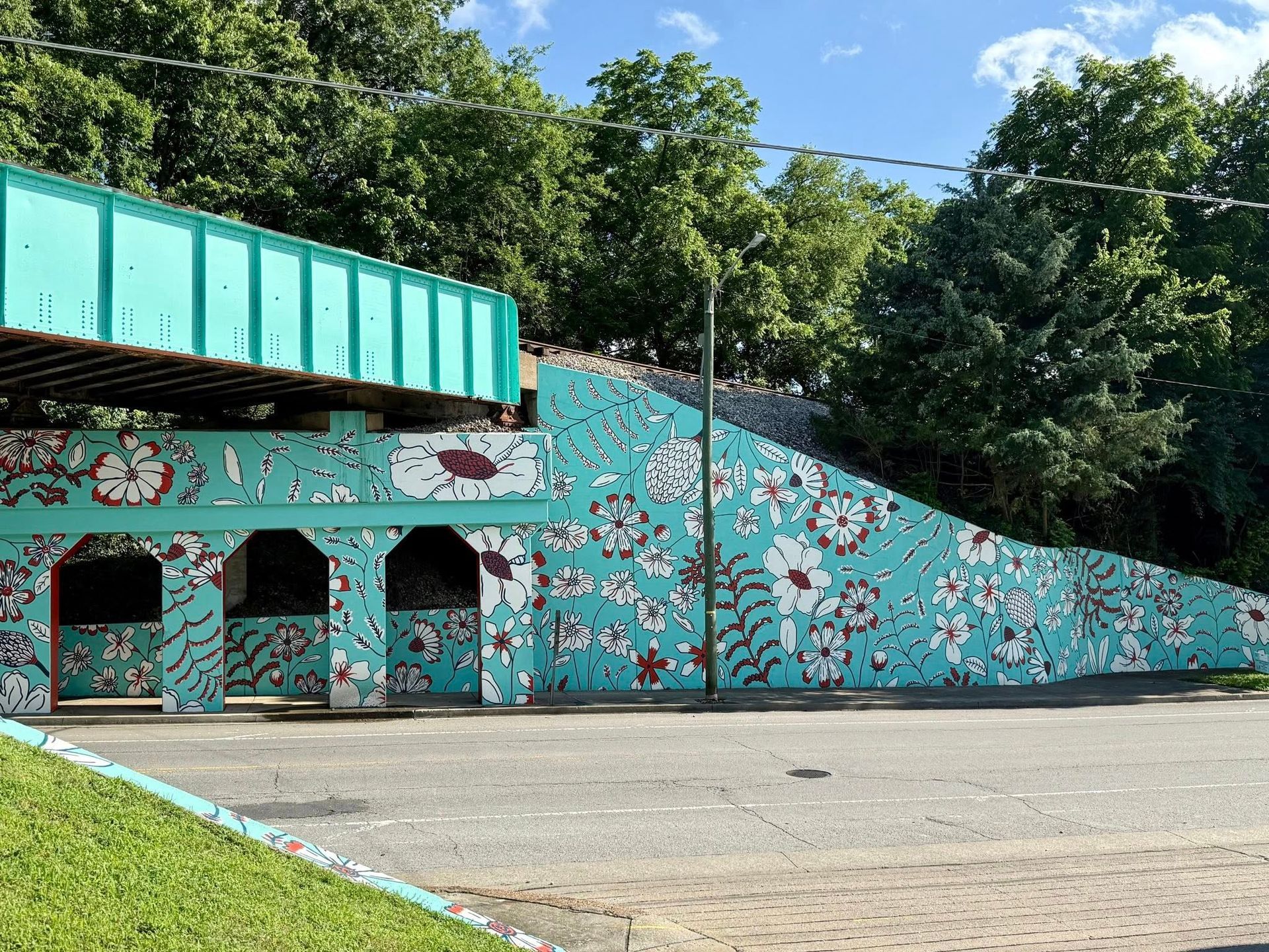 Turquoise mural on a bridge support with white and red floral patterns against a backdrop of trees.