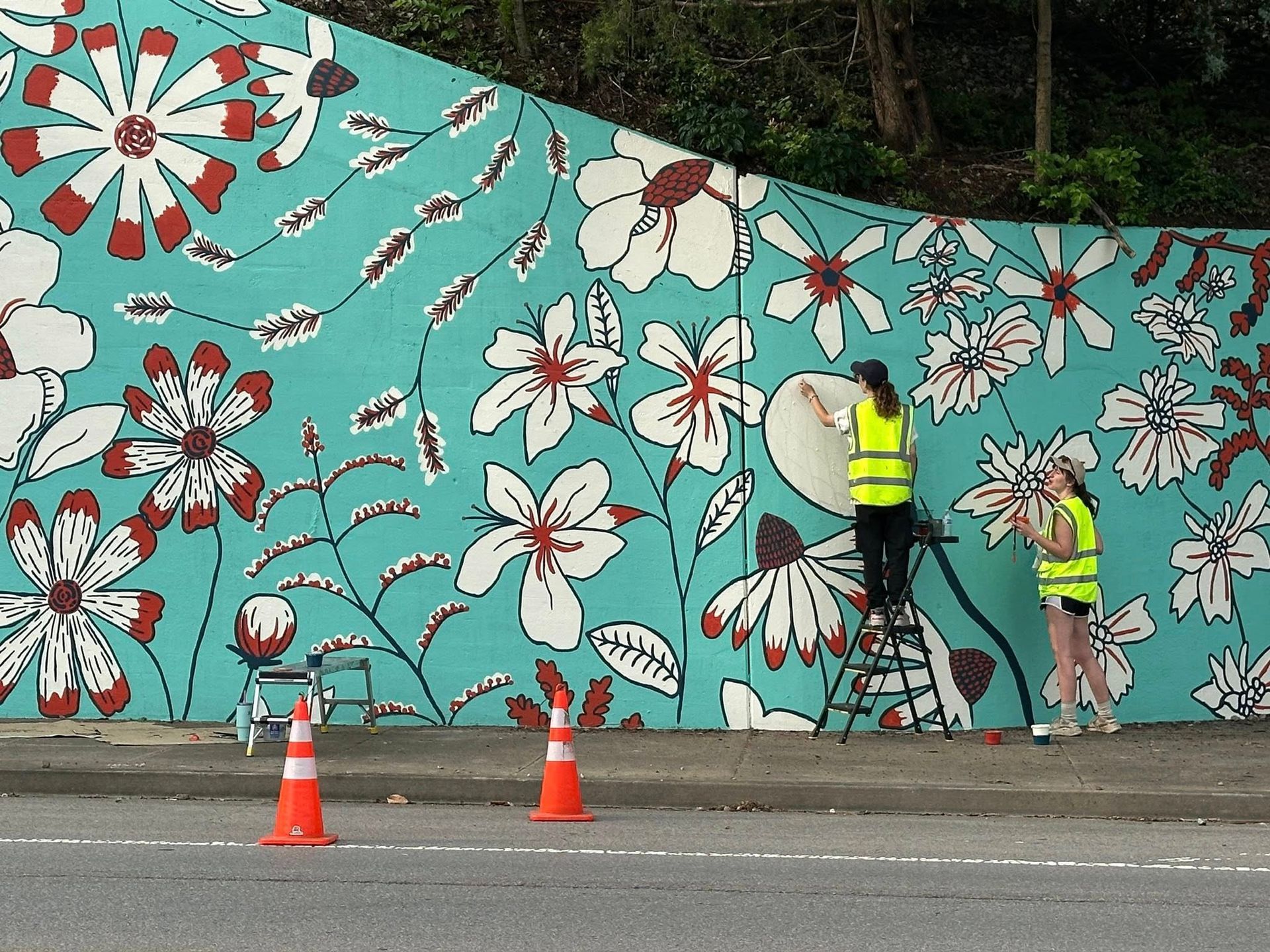 Two people painting a floral mural on a teal wall. One stands on a ladder, others on ground. Safety cones in foreground.
