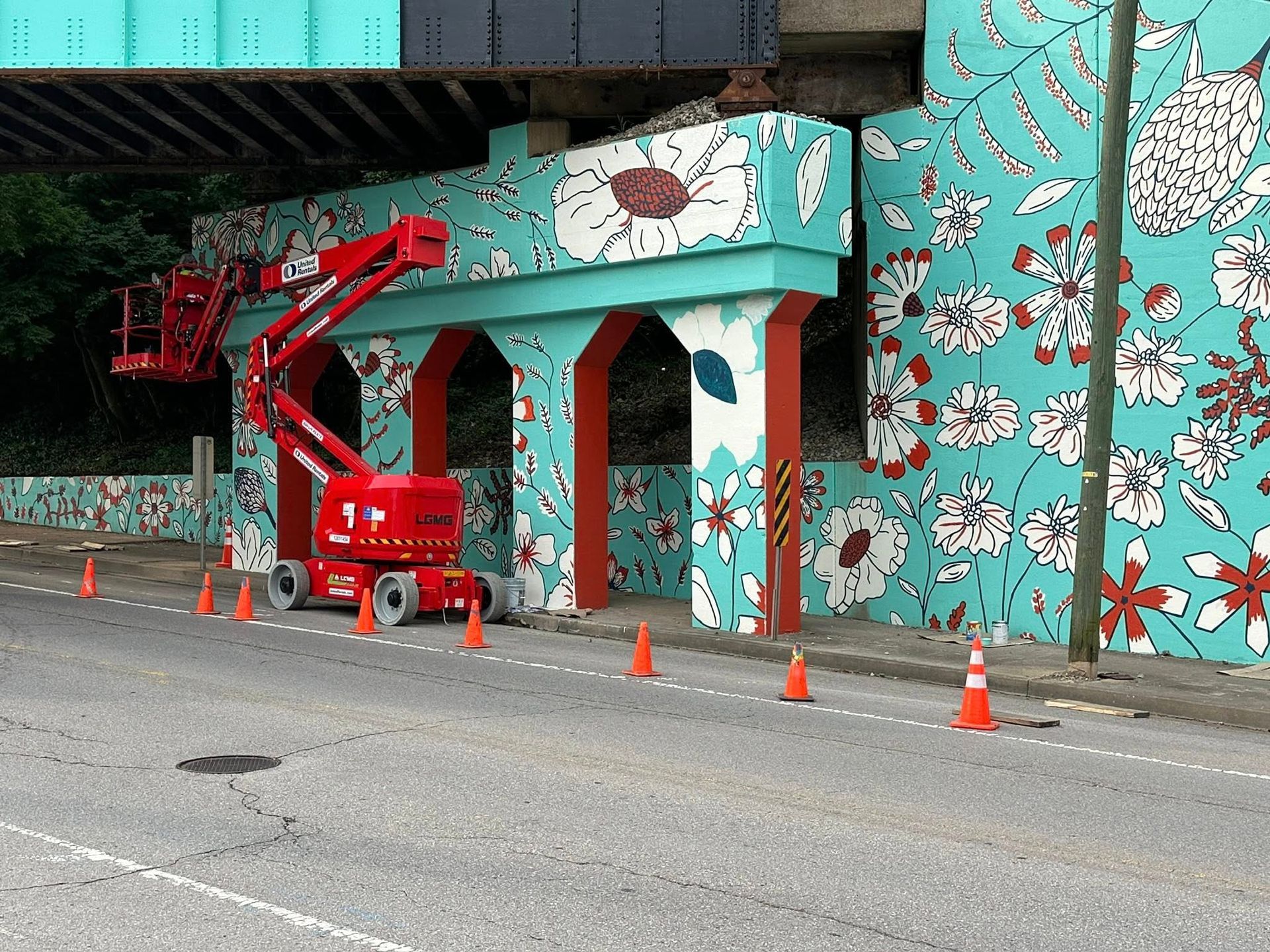 A red lift truck is parked under a bridge with teal and floral mural. Orange cones line the street.