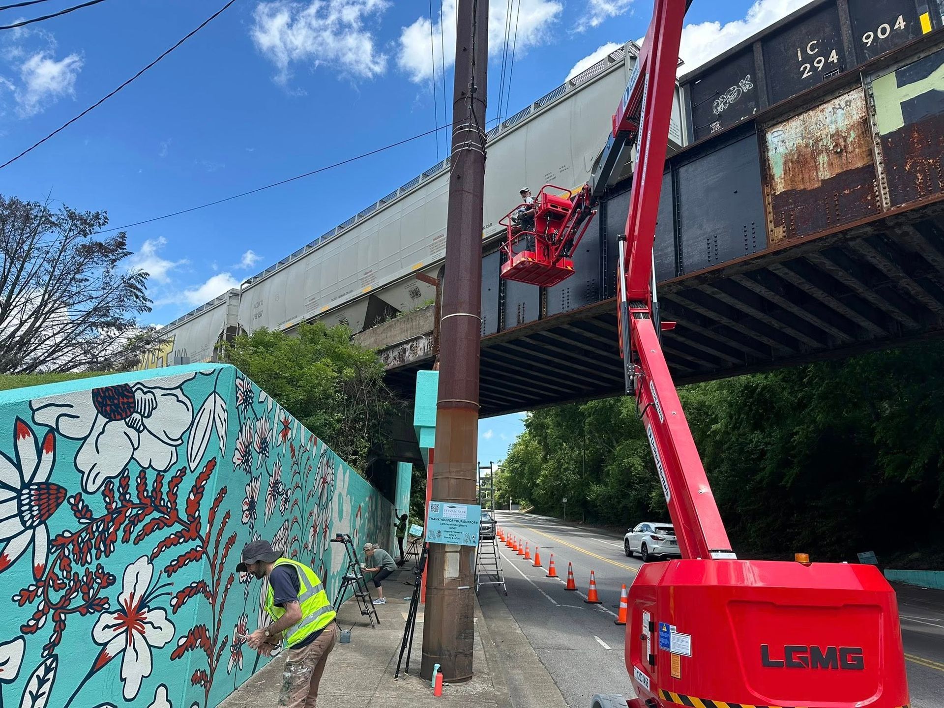 Mural artist paints on wall; bridge and workers in lift; power pole. Road closed with cones.