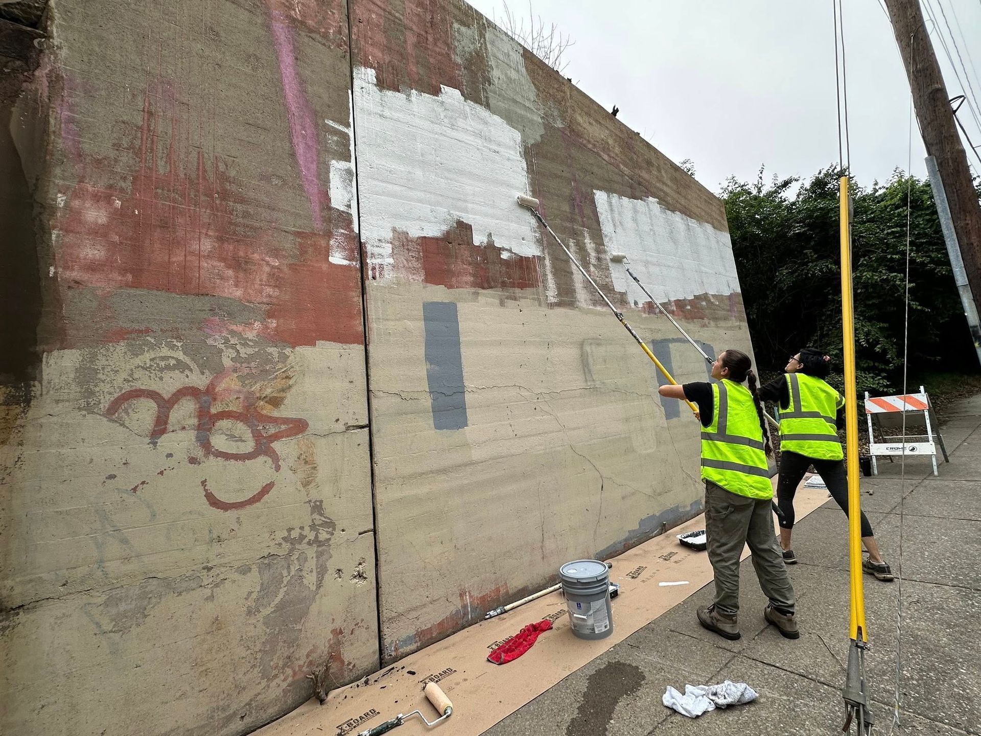 Two people in vests paint over graffiti on a concrete wall outdoors.
