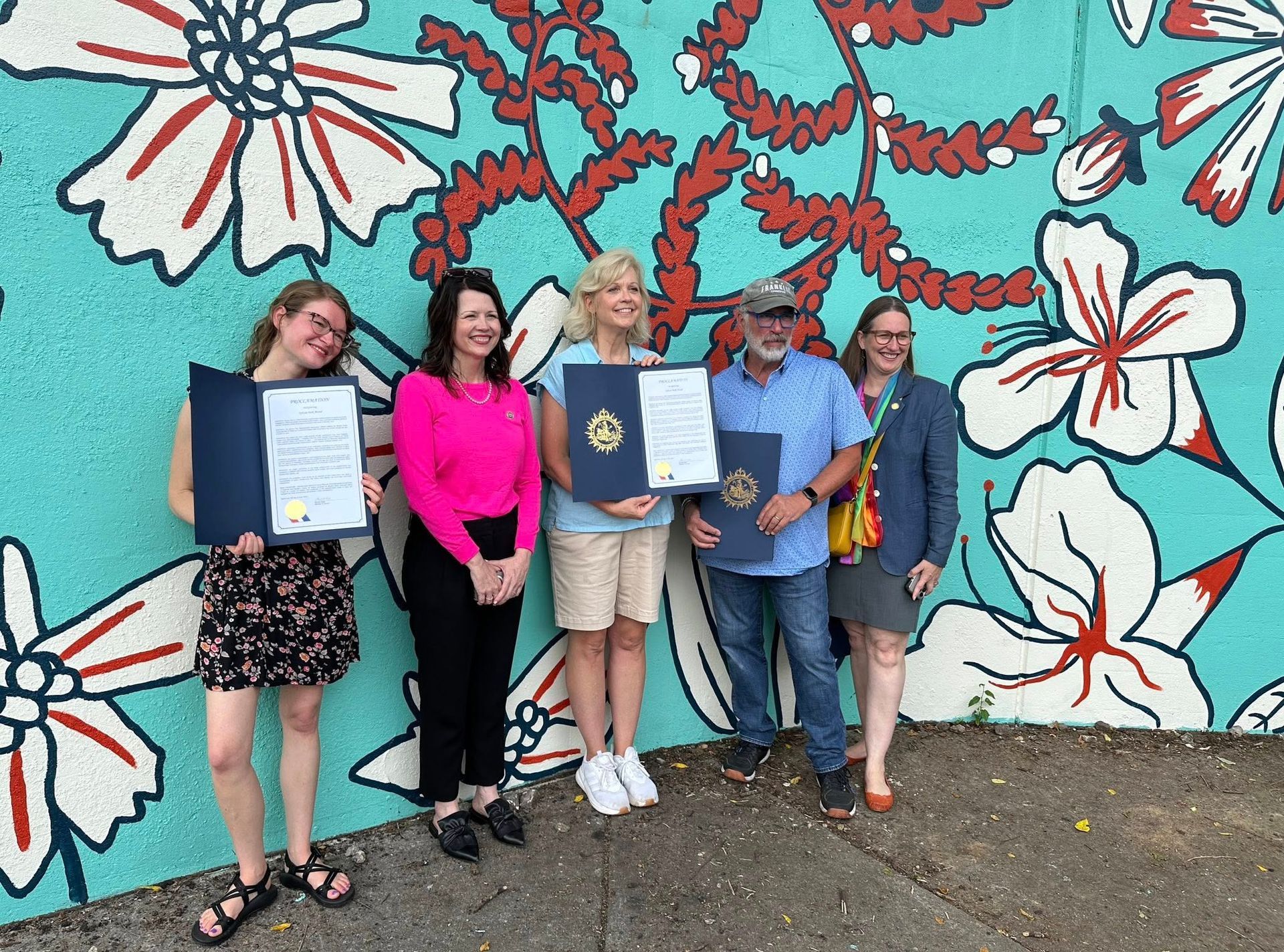 Five people stand in front of a colorful floral mural, holding documents.