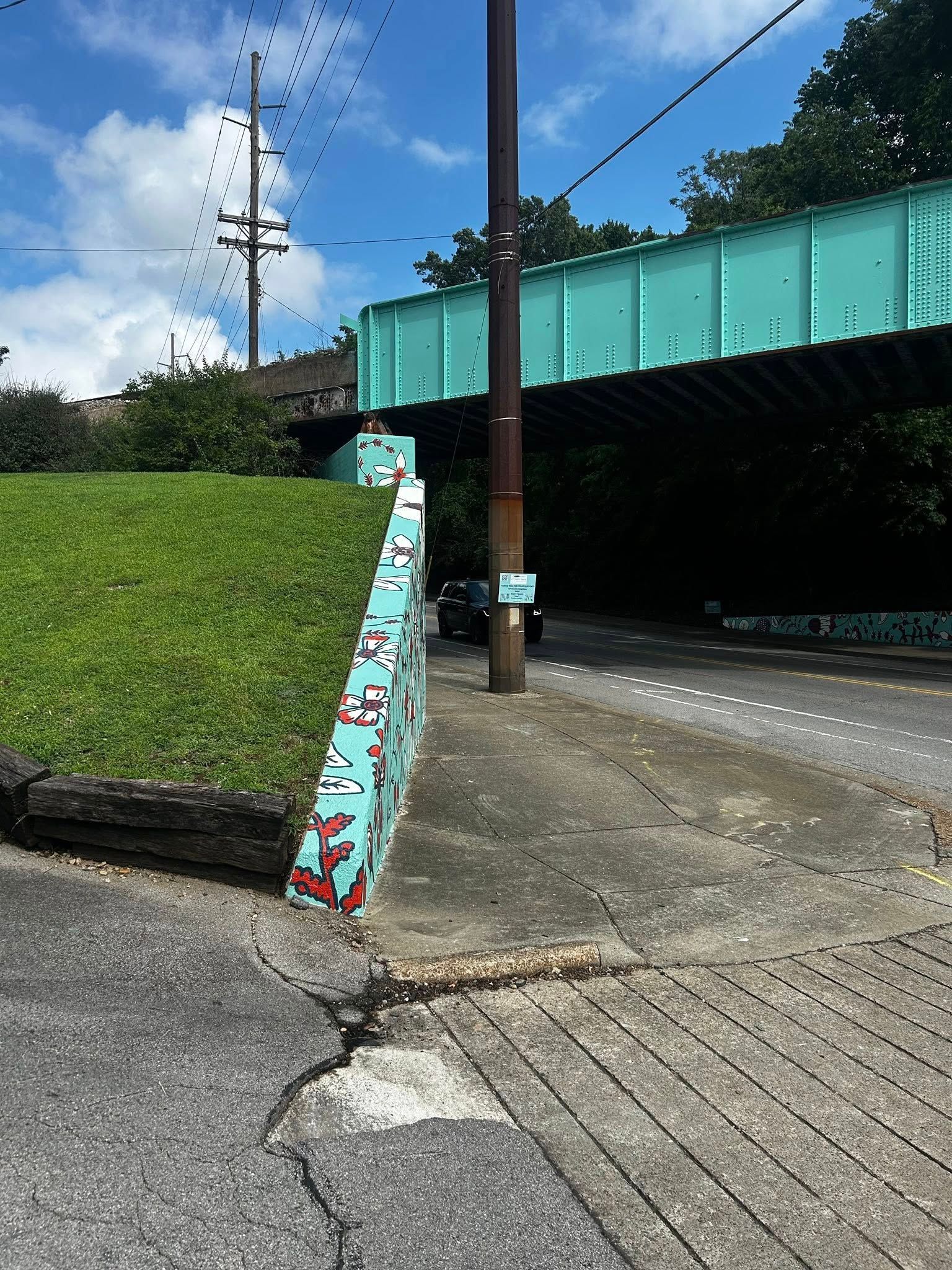 Green bridge over a road with a grassy hill on the left and a mural-covered support on the left.