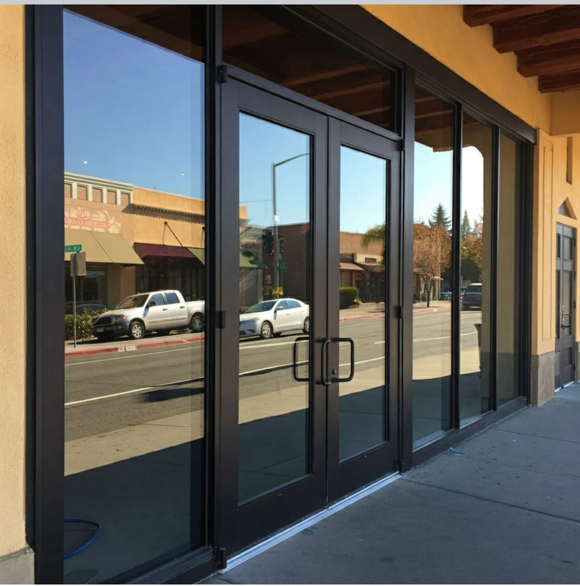 Glass storefront entrance with double black doors reflecting a sunny street and parked cars.