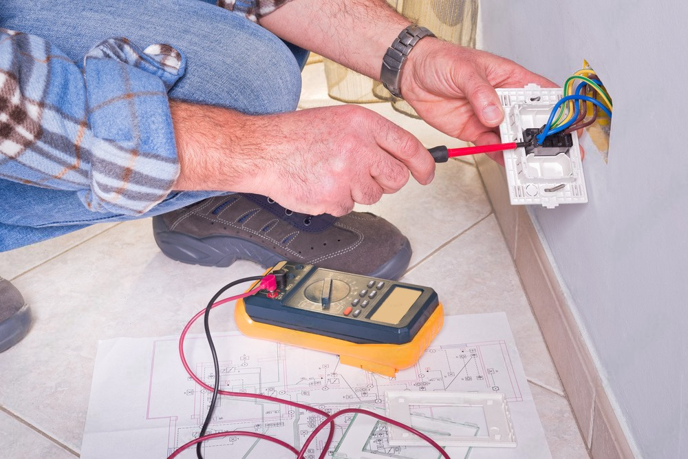 A man is working on an electrical outlet with a screwdriver and a multimeter.