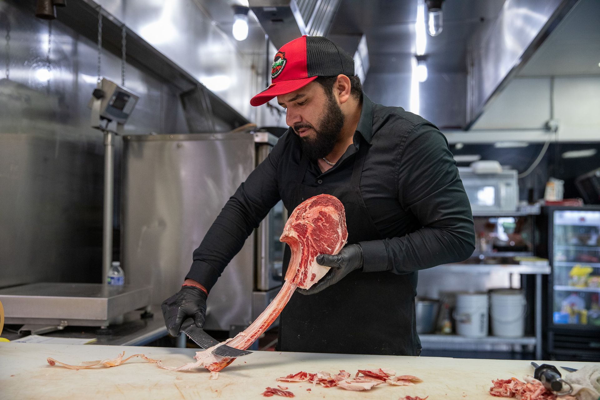 Butcher in black shirt and hat, wearing gloves, trims a large tomahawk steak in a restaurant kitchen.