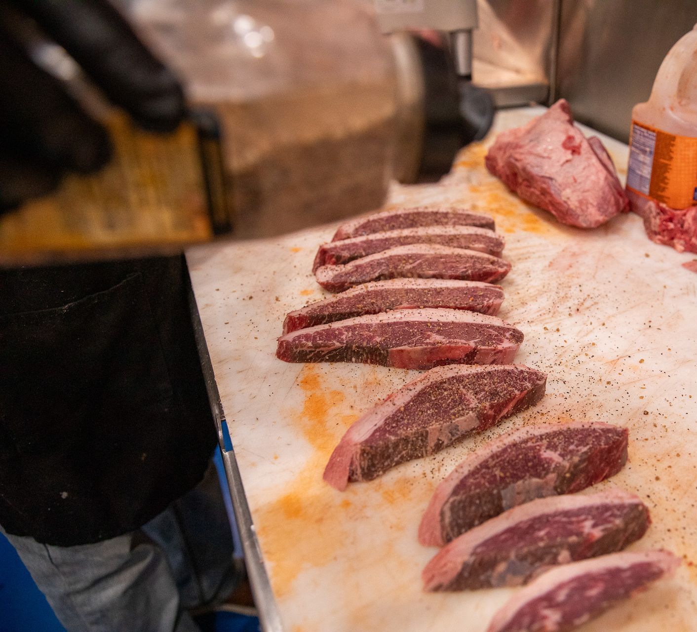 A person seasoning sliced raw steaks on a white cutting board with pepper, in a kitchen setting.