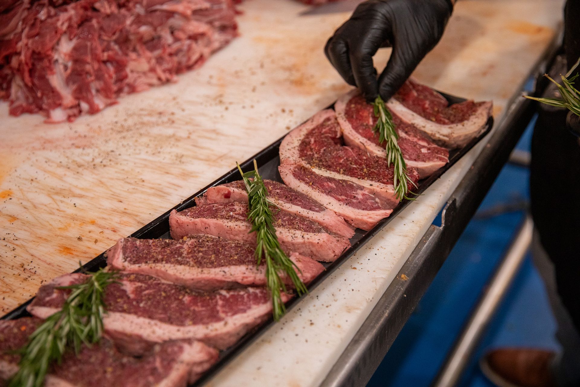 A butcher with black gloves garnishes seasoned steaks with rosemary sprigs on a white cutting board.
