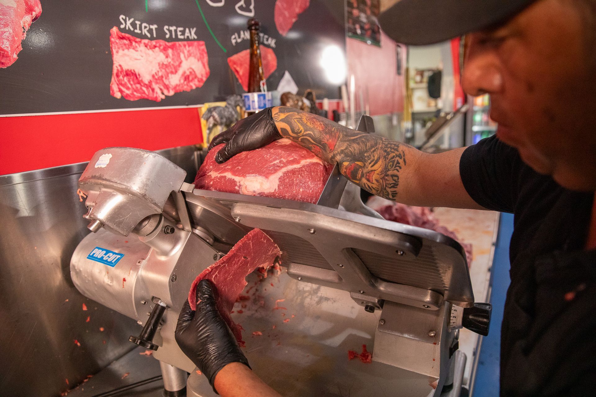 A butcher uses a meat slicer to cut a large piece of red meat. The butcher is wearing gloves and has tattoos on his arm.