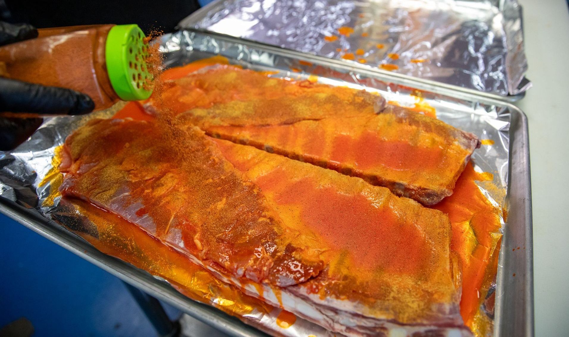 Ribs being seasoned with a red spice from a shaker on a foil-lined baking sheet.