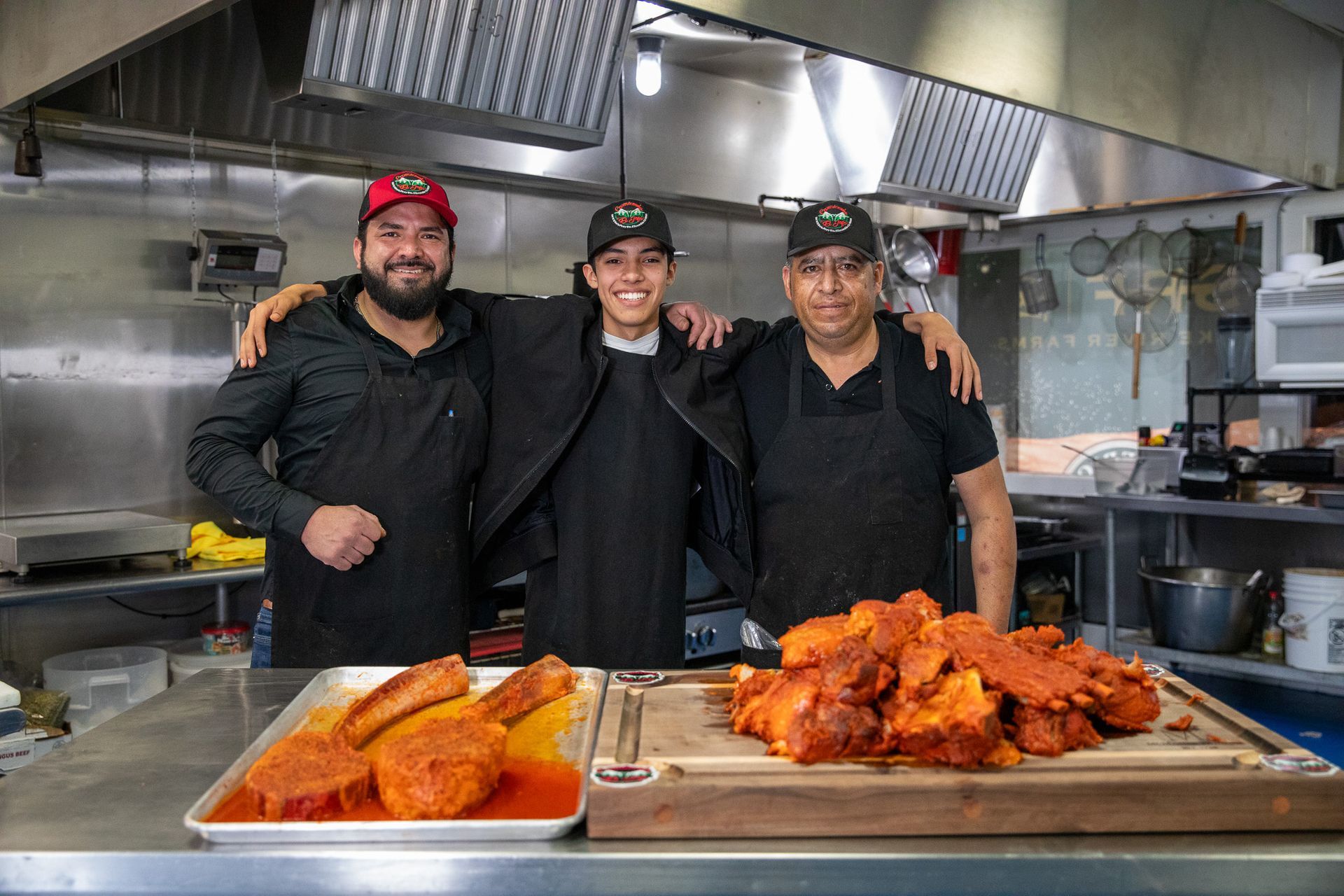 Three people in a commercial kitchen with trays of seasoned meat.