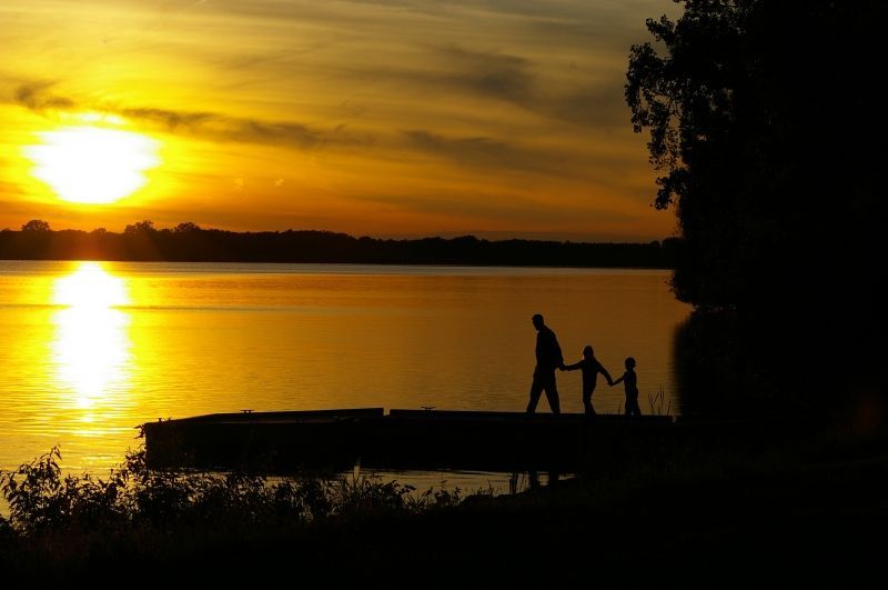 A man and two children are standing on a dock holding hands at sunset.