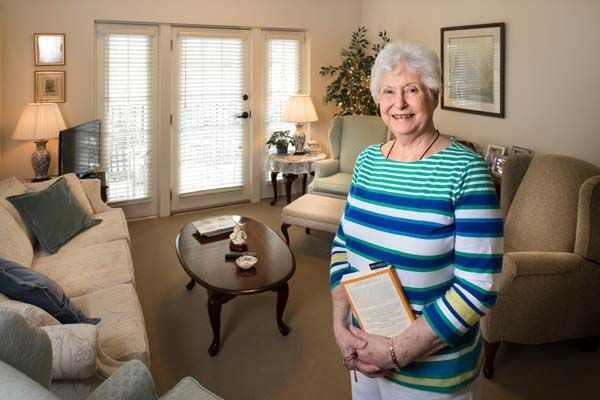 An elderly woman is standing in a living room holding a book.