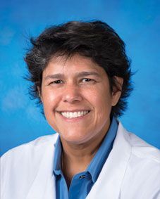 A woman in a white coat and blue shirt is smiling for the camera.