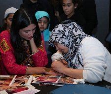 Two women are sitting at a table getting henna tattooed on their hands.