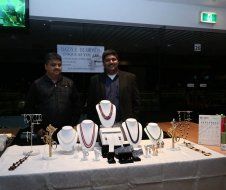 Two men are standing in front of a table with necklaces on it.