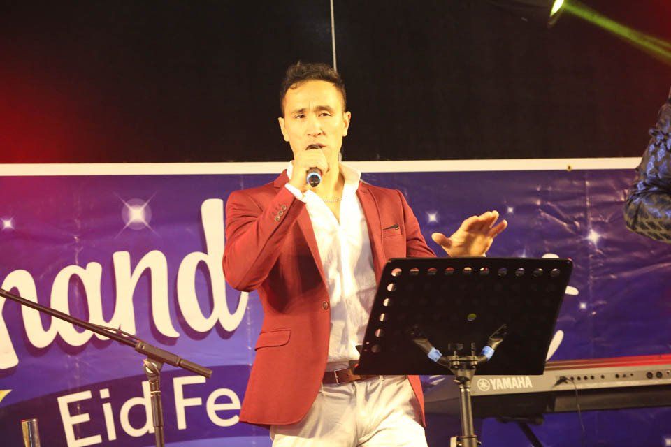 A man singing into a microphone in front of a sign that says grand eid fe