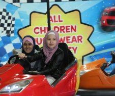 Two girls are riding a bumper car in front of a sign that says all children wear safety