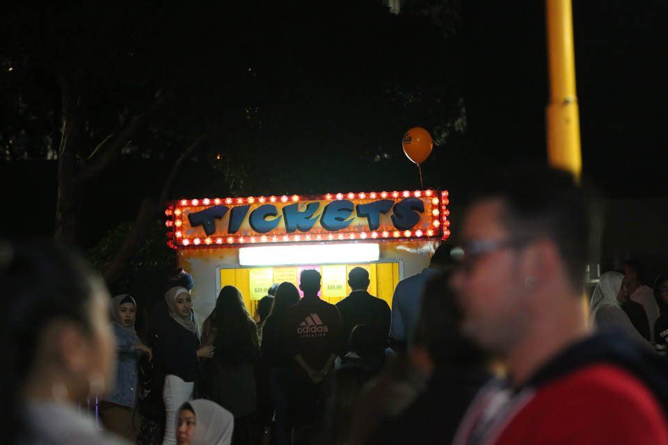 A group of people are standing in front of a tickets booth at a carnival.