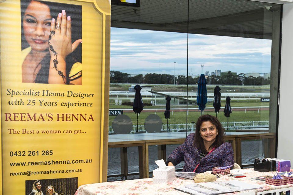 A woman is sitting at a table in front of a sign for reema 's henna