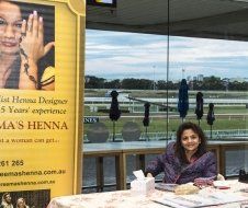 A woman is sitting at a table in front of a sign that says ma 's henna.