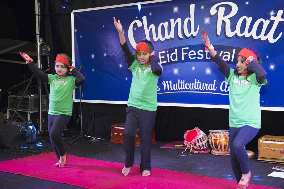 Three girls are dancing in front of a sign that says chand raat eid festival