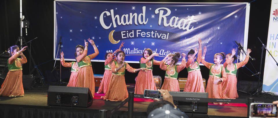 A group of people are dancing on a stage in front of a banner that says chand raat eid festival.