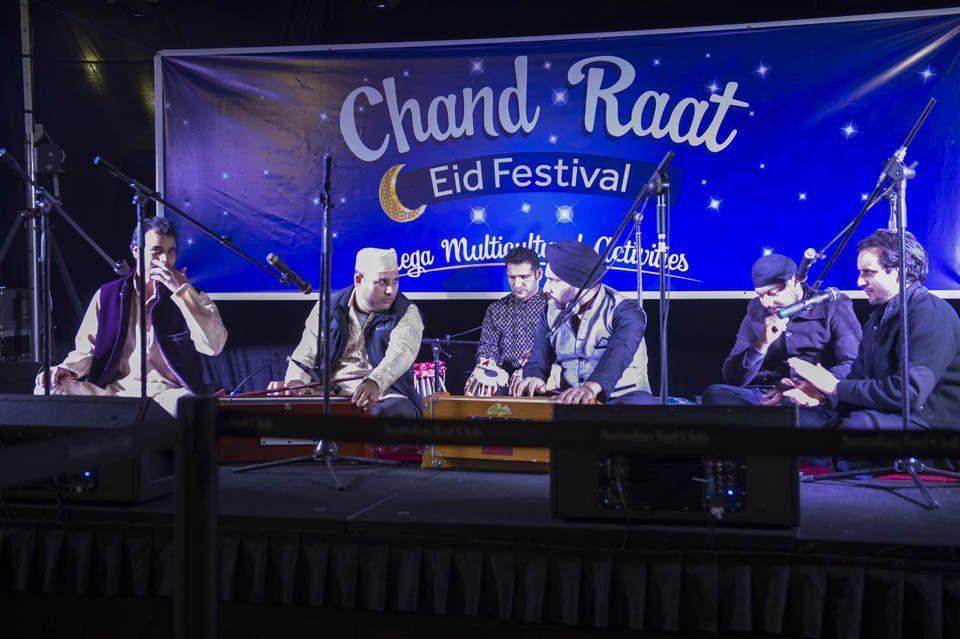 A group of men are playing instruments on a stage in front of a banner that says chand raat eid festival.