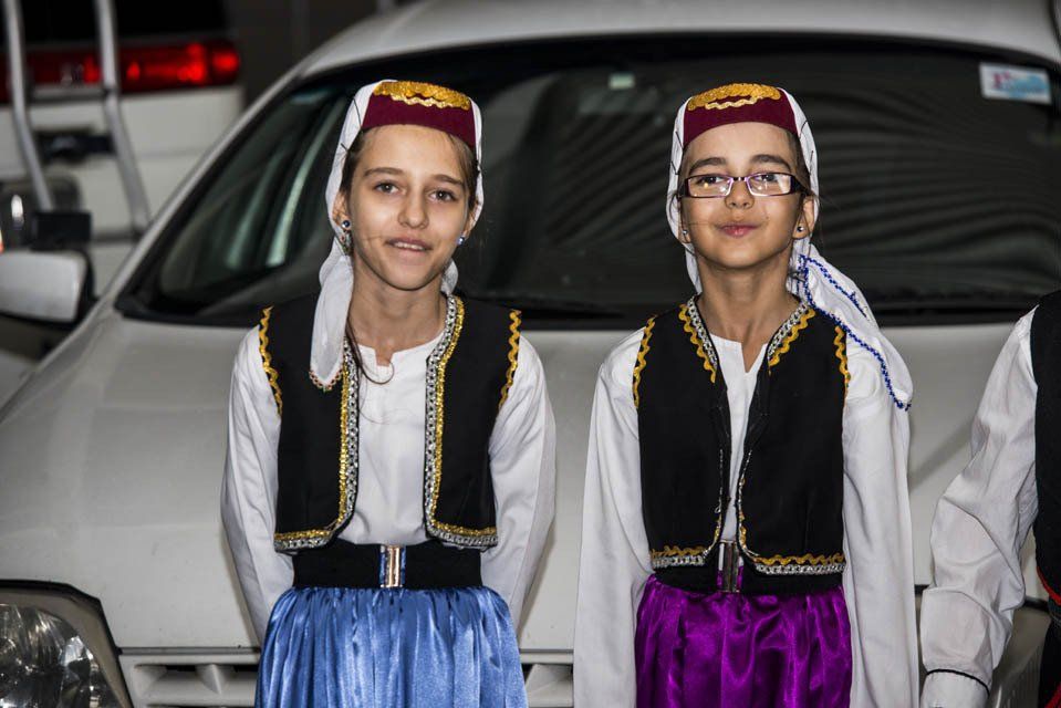 Two girls wearing traditional clothes are standing in front of a car