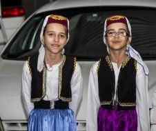 Two young girls wearing traditional clothes are standing next to each other in front of a car.