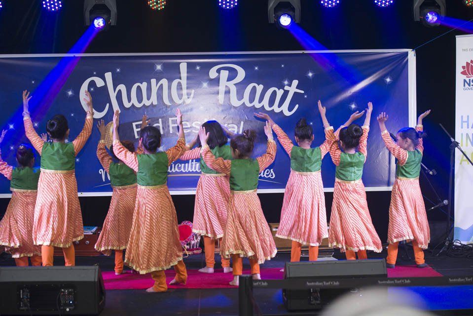 A group of young girls are dancing on a stage in front of a sign that says chand raat.