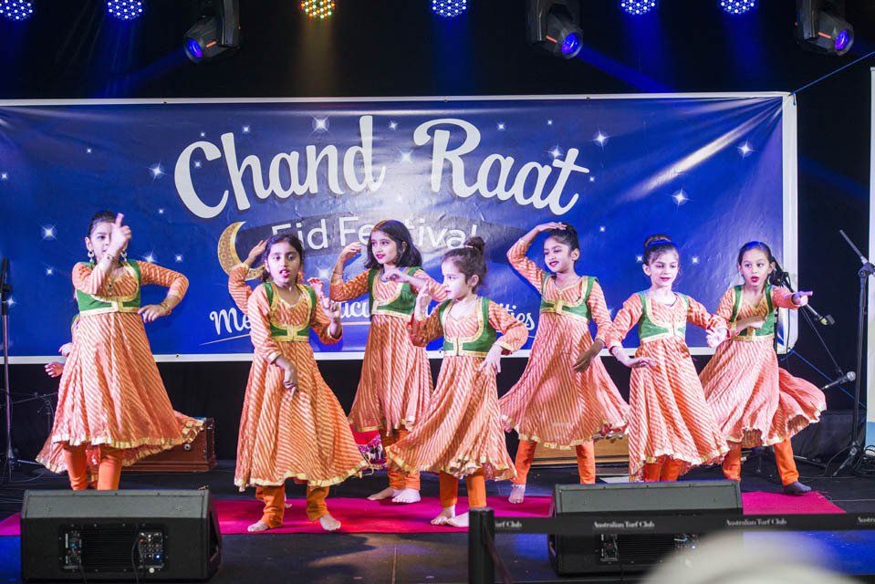 A group of young girls are dancing on a stage in front of a banner that says chand raat.