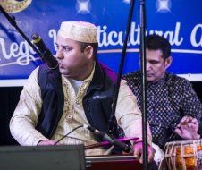 A man is sitting in front of a microphone while another man plays drums.