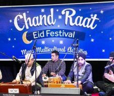A group of men are playing instruments on a stage in front of a banner for the chand raat eid festival.