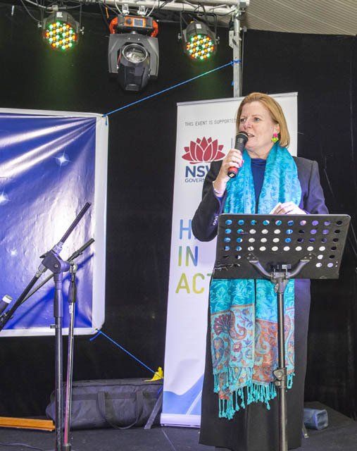 A woman stands at a podium holding a microphone in front of a sign that says nsw