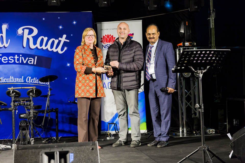 Three people are standing on a stage in front of a sign that says raat festival.