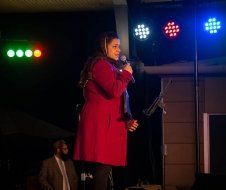 A woman in a red coat is standing on a stage holding a microphone.