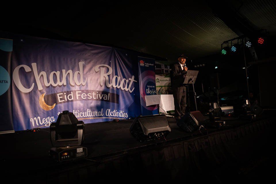 A man is standing on a stage in front of a banner that says chand road eid festival.
