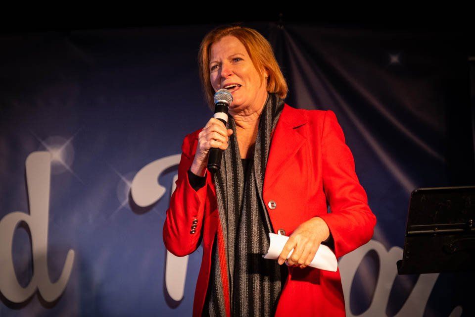 A woman in a red jacket is speaking into a microphone.