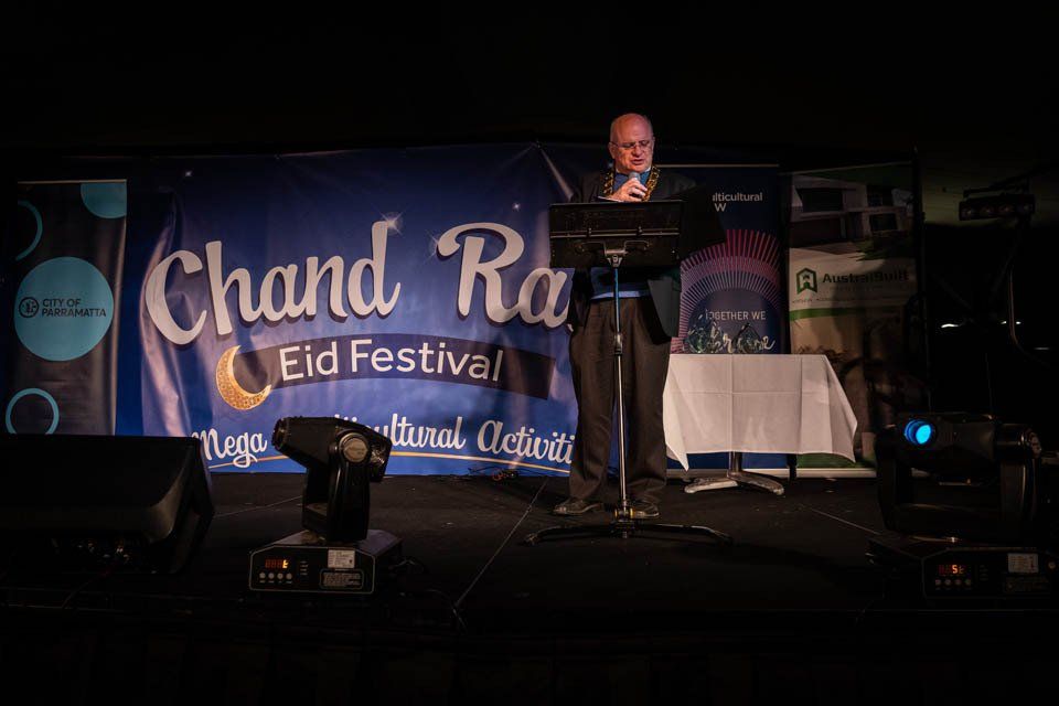 A man is standing on a stage in front of a banner that says chand ra eid festival.