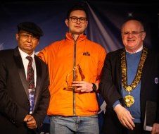 A man in an orange jacket is holding a trophy between two men in suits.