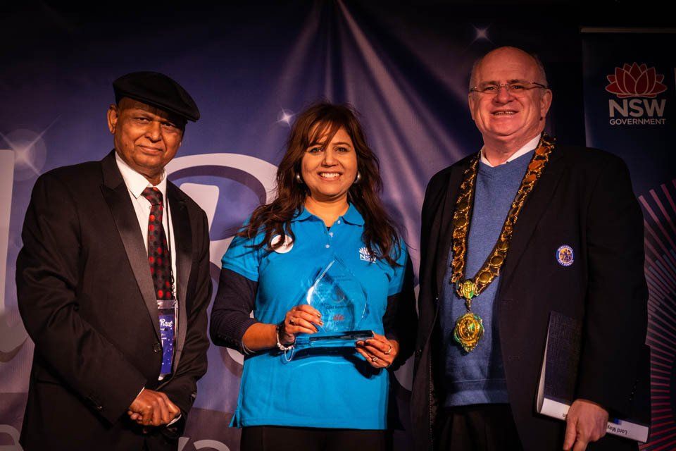 A woman in a blue shirt is holding a trophy between two men in suits.