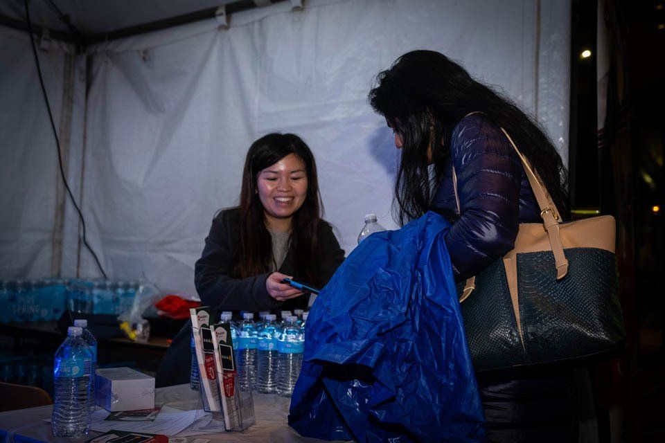 Two women are sitting at a table with bottles of water and a blue tarp.