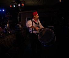 A man in a red hat and suspenders is playing drums in a dark room.